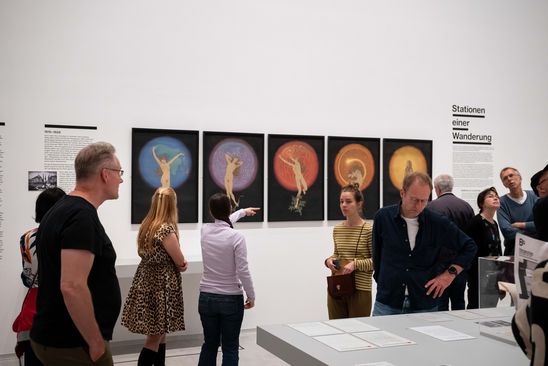 Photo: Some people stand in the provenance exhibition and look at works of art on the wall and archive material on a table in the room.