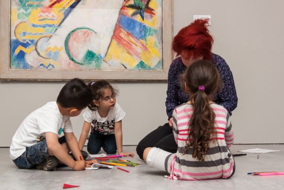 Foto: Kinder und eine erwachsene Person sitzen auf dem Boden im Ausstellungsraum. Auf dem Boden liegen viele Stifte.