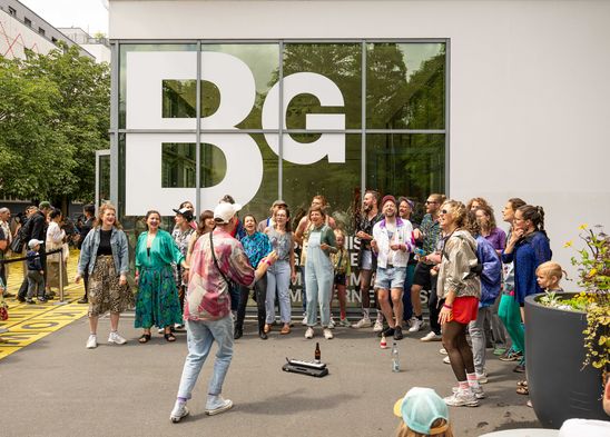 Photo: A group of colourfully dressed people stand in a semi-circle in front of the Berlinische Galerie and sing.