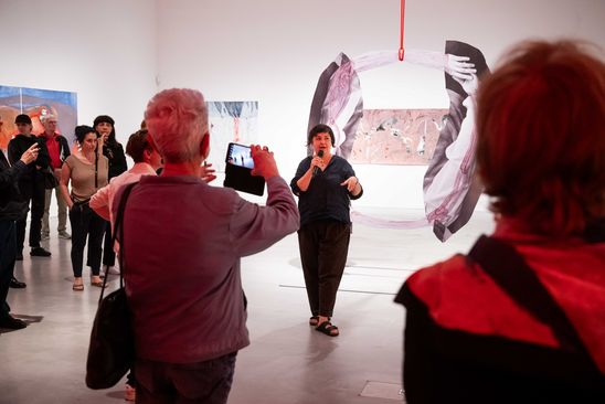 Photo: The artist during the tour of the exhibition space. Many participants are standing around her, some of them taking photos. Some works can be seen in the background.