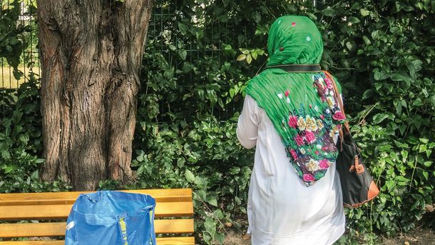 Foto: Eine Frau steht uns mit dem Rücken zugewandt neben einer Parkbank. Eine Kameraschlaufe hängt um ihren Hals und sie blickt auf Sträucher und Bäume.