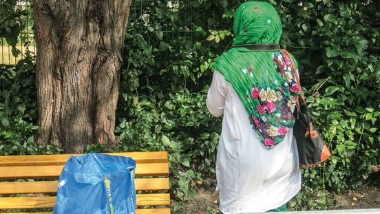 Foto: Eine Frau steht uns mit dem Rücken zugewandt neben einer Parkbank. Eine Kameraschlaufe hängt um ihren Hals und sie blickt auf Sträucher und Bäume.