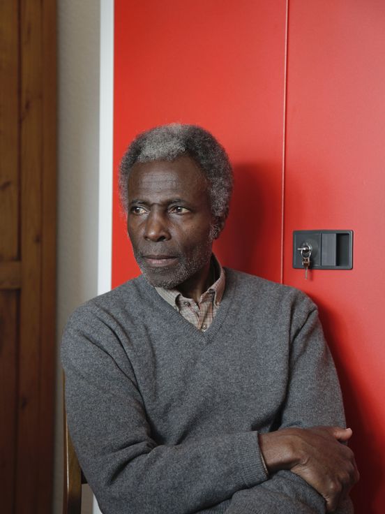 Portrait: Akinbode Akinbiyi steht in dunkelgrauem Pullover vor einem leuchtend roten Metrallschrank, den Blick leicht zur Seite gerichtet.