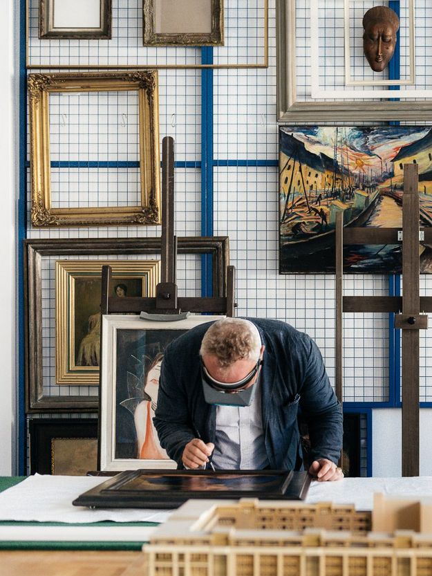 Foto: Person mit Vergrößerungsbrille betrachtet ein Kunstwerk mit Pinsel in der Hand. Dahinter an der Wand Rahmen und Kunstwerke.