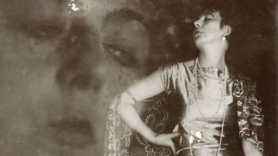 Fotografie: Die Fotografie zeigt die Künstlerin in einem langen Satin-Kleid mit langer, oranmentaler Schleppe und in Pose. Im Hintergrund ist leicht transparent eine Nahaufnahme ihres Gesichts zu sehen.