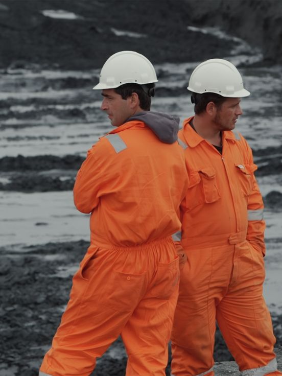 Filmstill: Zwei Personen in leuchtend-oranger Arbeitskleidung und weißen Helmen stehen vor einer Baugrube, Kopf und Körper jeweils in eine andere Richtung gewandt.