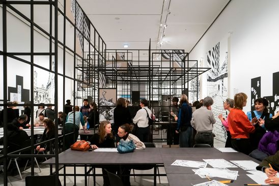 Foto: Blick in den Ausstellungsraum von „Kotti-Shop/SuperFuture" mit vielen Menschen, zwei langen Tischen und einer großen Gitterkonstruktion.