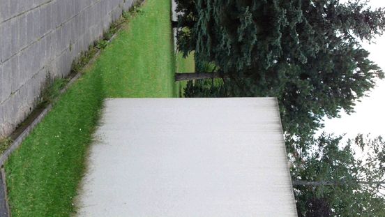 Filmstill: Blick auf einen gepflasterten Weg, eine von Gras umgebene weiße Mauer und dahinter Bäume. Das Bild ist eigentlich ein Hochformat, in der Ansicht aber einmal nach rechts gedreht.