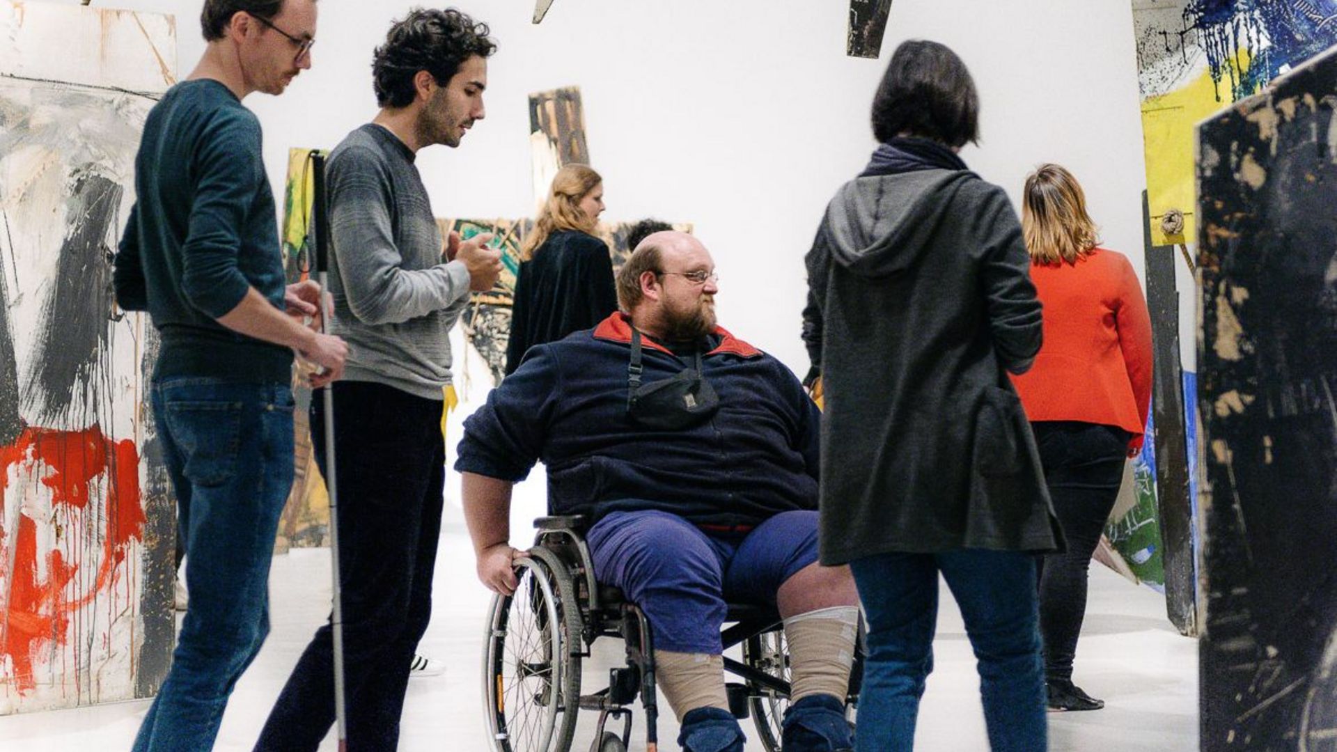 Foto: Menschen bewegen sich in einer Rauminstallation. Eine Person mit Rollstuhl, eine Person mit Blindenlangstock.