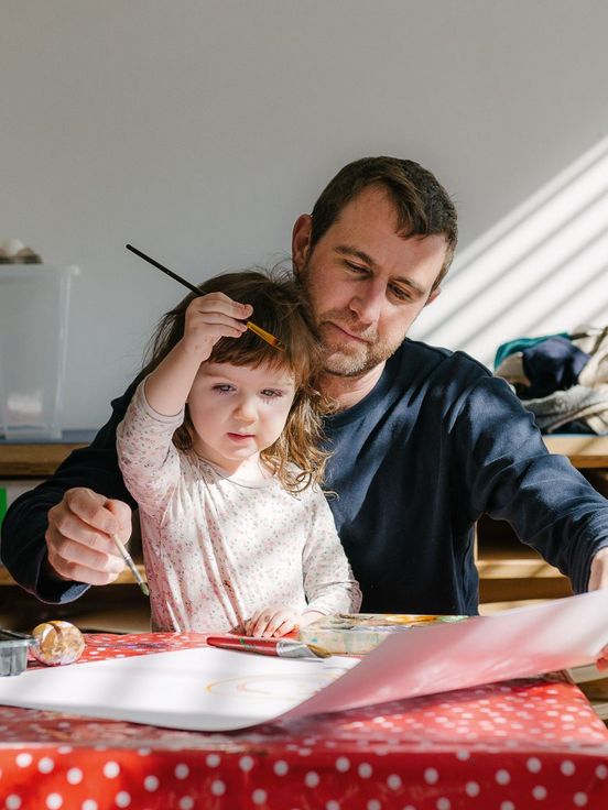 Foto: Erwachsene Person und Kind sitzen an einem Tisch. Sie malen gemeinsam ein Bild.