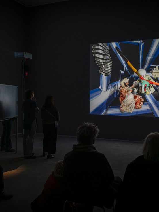 Foto: Ein paar Menschen sitzen und stehen in einem Videoraum und schauen auf eine große Leinwand.