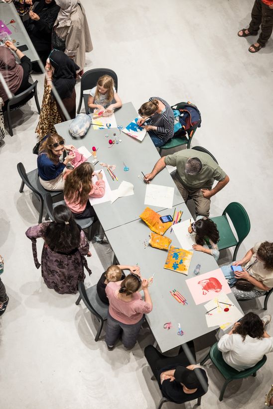 Photo: View from above of a long table at which many people are working creatively.