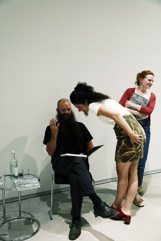 One person is sitting on a chair and signing a catalogue while two other people are standing next to her.