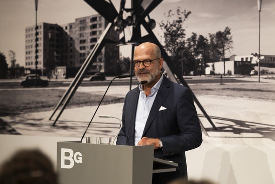 Foto: Thomas Köhler steht an einem Redner*innen-Pult vor einer Fototapete in schwarz-weiß. Die Tapete zeigt eine Skulptur von Hans Uhlmann am Hansaplatz in Berlin.
