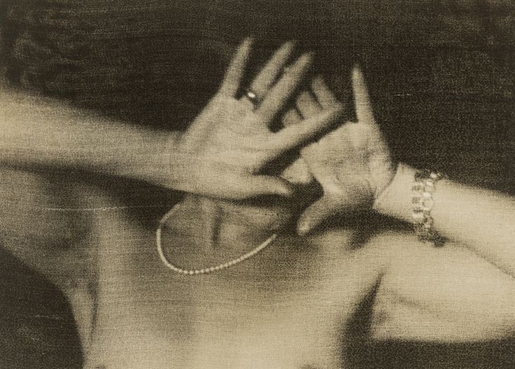Körnige Schwarz-Weiß-Fotografie: Porträt einer unbekleideten Person mit Perlenkette, die beide Hände vor das Gesicht hält, sodass dieses nicht sichtbar ist. Die Hände tragen Schmuck, darunter ein Ring und ein Armband. 