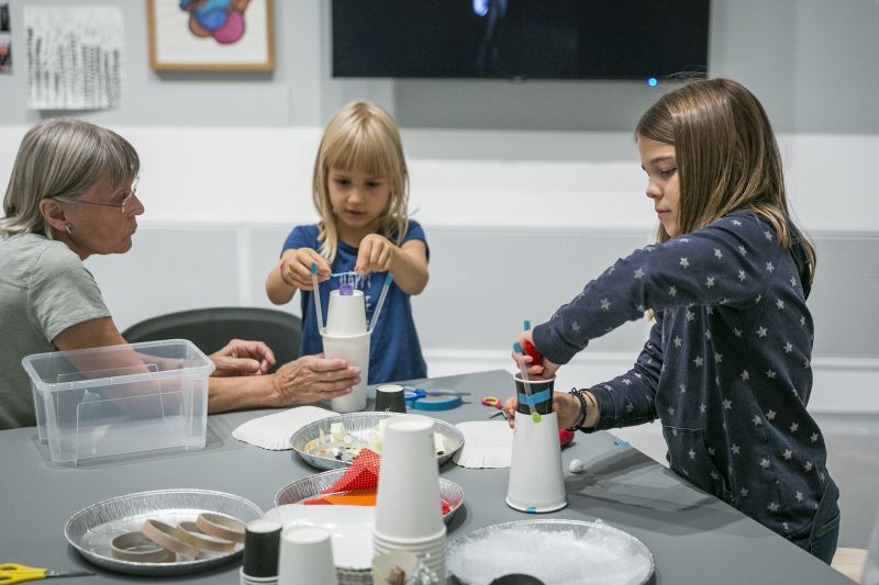 Berlinische-Galerie-family-day Photo: Two children and an adult are sitting around a table with handicraft materials. The children are making things out of paper cups.
